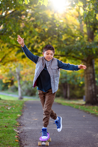 Elementary age multiracial boy has fun skateboarding on a paved walking path through a public park on a beautiful Autumn day.