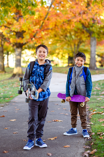Portrait of two elementary age Eurasian boys wearing backpacks and carrying skateboards smiling directly at the camera while walking to school on a paved walking path through a public park in their residential neighborhood on a beautiful Autumn day.