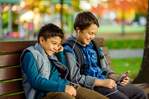 Elementary age Eurasian brothers sit together on a park bench and share a pair of headphones while watching a video on a smart phone.