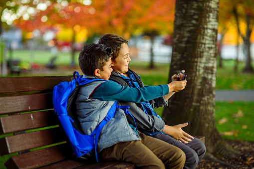 Two Eurasian elementary age boys sit on a park bench after the first day of school and take a selfie on a smart phone before walking home.