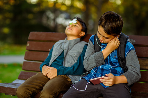 An elementary age boy uses his smart phone for social media and gaming while his younger brother entertains himself with a fidget spinner as they relax on a bench in a public park after school