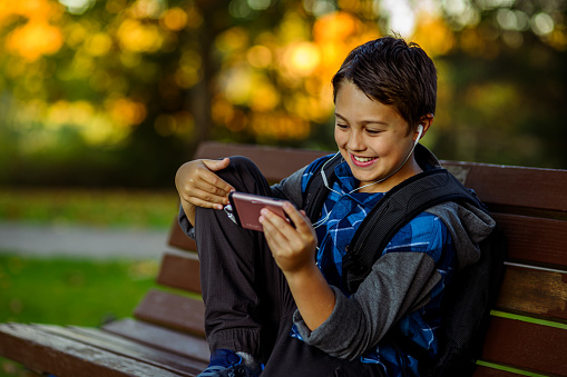 An cheerful multiracial elementary age boy uses his smart phone for social media and gaming while sitting on a park bench after school.