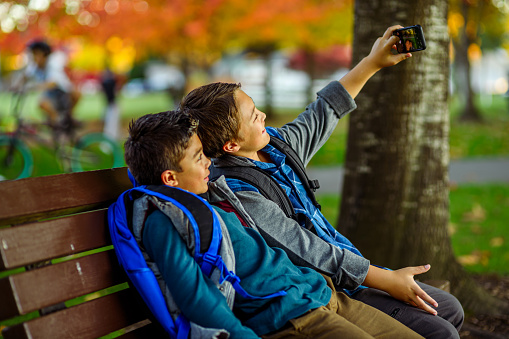 Two Eurasian elementary age boys sit on a park bench after the first day of school and take a selfie on a smart phone before walking home.