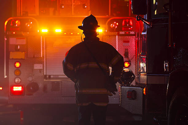 Firefighter Firefighter silhouetted against a fire truck with flashing lights at an emergency scene. extinguishing stock pictures, royalty-free photos & images