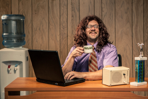 Silly retro office scene of a long haired shaggy looking dude working at his desk. He is holding a mug filled with coffee or water. Water cooler in background and copy space available.