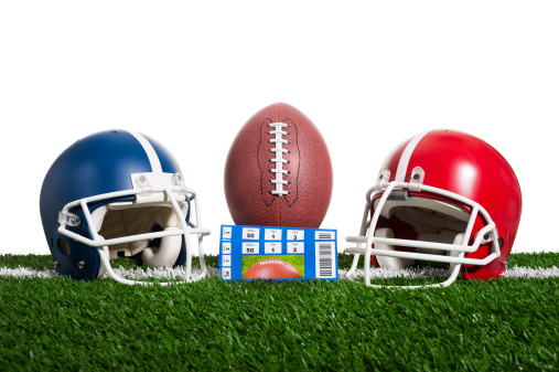 American Football - Tickets, A low angle view of a pair of ticket stubs in front of a football sitting on a tee over a yard line against artificial turf with a blue and red helmet flanking them