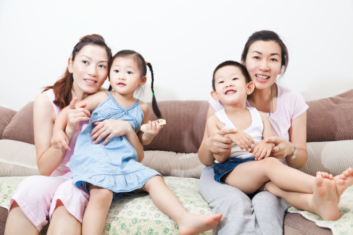 Happy east asian mothers and children sitting on sofa at home watching TV together
