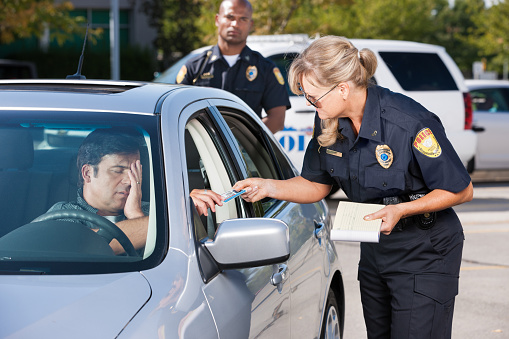 A mature female law enforcement officer stands by a vehicle she has stopped and takes the driver's license from a middle aged man while her partner stands by in the background.