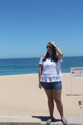 A woman looking to the left, wearing a white blouse, shorts and sunglasses. The beach in the background
