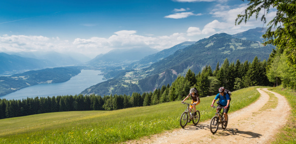 A couple is biking uphill on a scenic forest road with an overview of Lake Millstatt, Carinthia, southern Austria. The region is a very attractive holiday area and is famous for hiking, mountainbiking and swimming during summertime. In wintertime there lots of skiing areas as well.Canon EOS 5D Mark III, 1/250, f/9, 24 mm.More pictures of mountainbike actions are available in my LB 