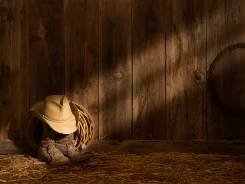 This images shows sun streaks across a barn wood wall onto a pair of drover packer boots, straw hat and lasso. The barn floor is littered with hay and a coil of dimly lit barbed wire adorns the wall. Image can be cropped to a wide panorama.