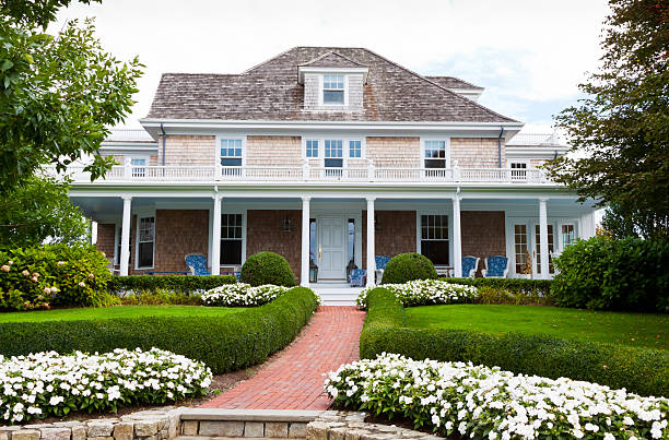 A view of a luxurious house in New England Luxury Home in Chatham, Cape Cod, Massachusetts, USA.  Beautifully landscaped front yard with flowers, lush grass and bushes is in foreground. Chairs are on the porch. Trees frame the house on both sides. cape cod photos stock pictures, royalty-free photos & images
