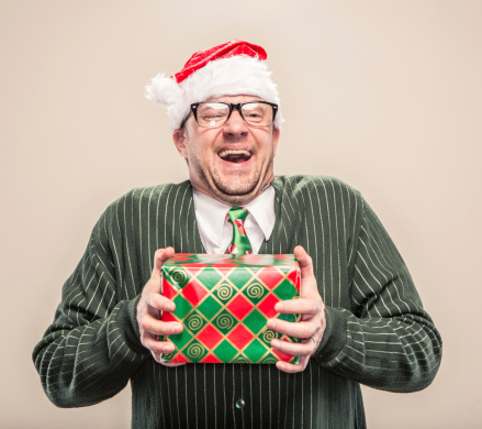A dorky nerd man in cardigan sweater holding holiday christmas gift. He is wearing a santa hat and a holiday festive neck tie.