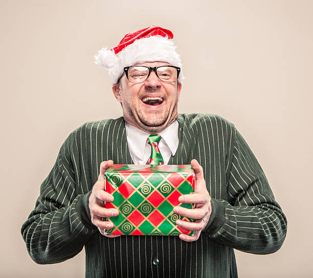 Nerdy Geek Christmas Man holding wrapped holiday gift A dorky nerd man in cardigan sweater holding holiday christmas gift. He is wearing a santa hat and a holiday festive neck tie. nerd sweater stock pictures, royalty-free photos & images