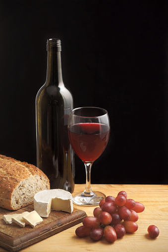 Still life with a bottle and glass of red wine, goat's cheese, red grapes and wholegrain bread . Photographed on a cutting board with a knife and a black background. Focus on the cheese.