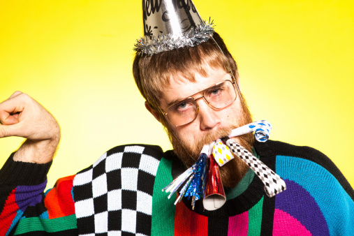 A nerdy bearded man with tons of party whistles and blowers in his mouth, party hat, and bright but super ugly sweater. Shot on a solid yellow background.