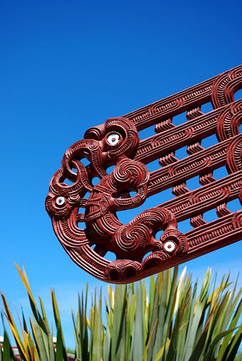 A Close-Up of a Carving on a Marae against a Blue Sky with Harakeke (NZ Flax) along the bottom.