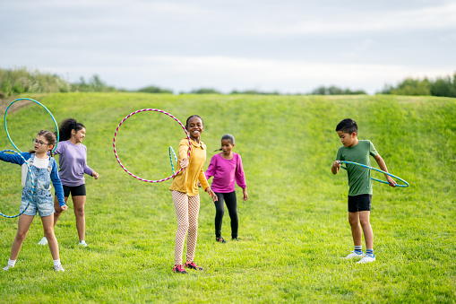 A small group of multi-ethnic school aged children are seen playing with Hula-Hoops during a recess break.  They are each dressed casually and are smiling as they focus on seeing who can spin their hoop the longest.