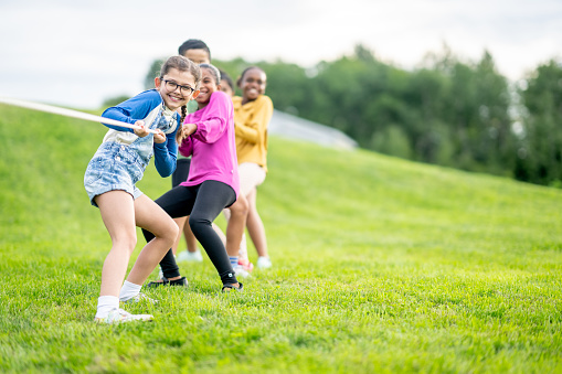 A small group of multi-ethnic school aged children are seen playing a game of Tug-of-War during a recess break.  They are each dressed casually and are smiling as they focus on pulling their team to a win!