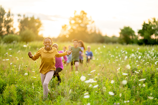 A small group of school aged children are seen walking through a field of tall grass as they take a walk and explore together.  They are each dressed casually and are smiling as they enjoy the warm summer air.