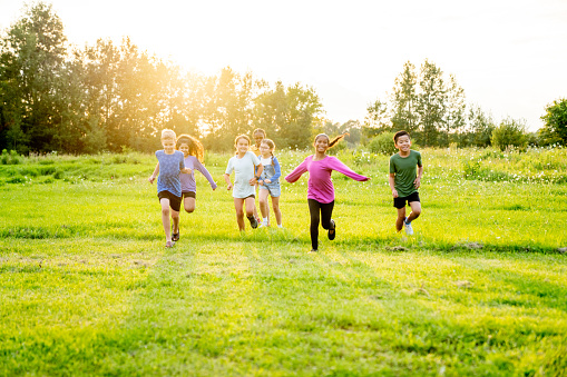 A small group of school aged children are seen running across the grass as they play at recess.  They are each dressed casually and are smiling as they enjoy the fresh air.