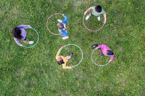 A small group of multi-ethnic school aged children are seen playing with Hula-Hoops during a recess break.  They are each dressed casually and are smiling as they focus on seeing who can spin their hoop the longest in this aerial view.