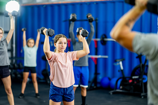 A small group of children work out together in a gym with free weights, under the direction of their fitness instructor.  They are each dressed comfortably in athletic wear and are focused on proper form and breathing.