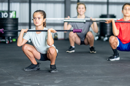 A small group of children work out together in a gym with barbells, under the direction of their fitness instructor.  They are each dressed comfortably in athletic wear and are focused on proper form and breathing.