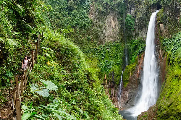 Woman walking along a nature trail in front of towering tropical waterfall in Costa Rica. Waterfall plunges 300 feet into the crater of an extinct volcano.