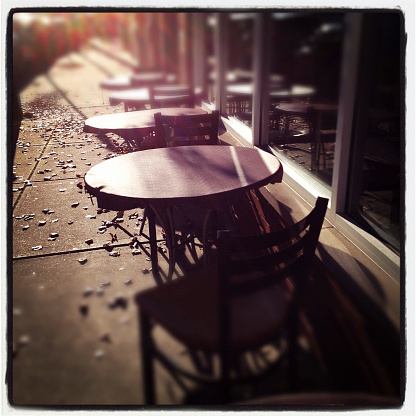 Outdoor Cafe. The photo is square, with a thick white border, and has had the color balance modified through the use of a filter via a popular image posting website. The photo is focused on a table with two chairs, and behind those are tables an chairs in a similar set up.