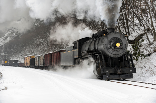 Freight train rounding curve through snowy rock cut beside a river with steam locomotive billowing smoke in the cold winter air like the Polar Express, historical reenactment in Cumberland, Maryland, MD, USA.