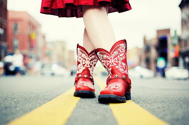 Cowgirl with Red Boots in the Road A cowgirl in red cowboy boots and dress has her legs crossed, standing on the double yellow line in the middle of a city street. Ground level view of the boots and legs. Taken in Nashville, Tennessee, USA. cowboy boot stock pictures, royalty-free photos & images