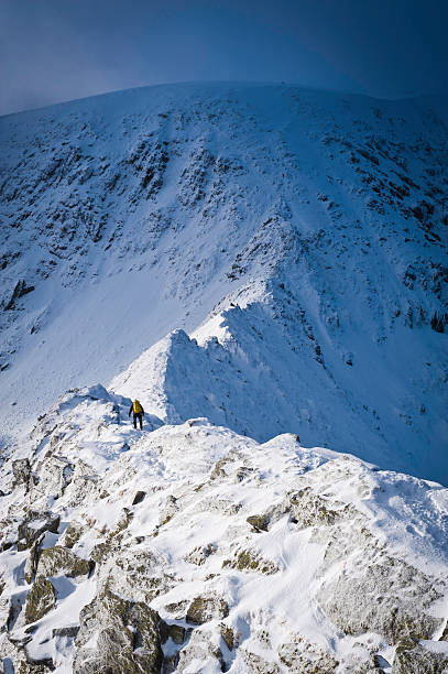 Mountaineer crossing snowy Striding Edge winter Lake District Cumbria UK Mountaineer climbing along the famous dramatic mountain ridge covered in full winter snows to the summit of Helvellyn above, Lake District National Park, Cumbria, UK. ProPhoto RGB profile for maximum color fidelity and gamut. striding edge stock pictures, royalty-free photos & images