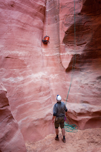 a man exploring a slot canyon looks up while his backpack is lowered down a sandstone cliff wall.  such dramatic adventure and exploration can be found in larry's canyon in the robber's roost zone of the san rafael desert, utah.  vertical wide angle composition.