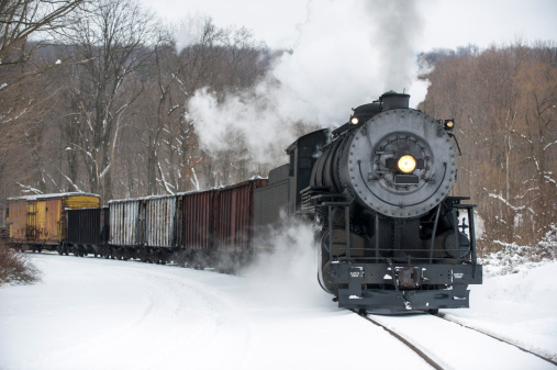 Steam locomotive pulling freight train rounding curve in winter snow pushing smoke, historical reenactment near Cumberland, Maryland, MD, USA.