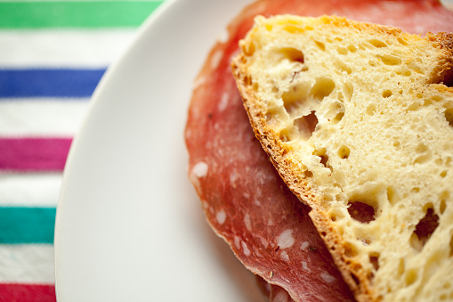 Horizontal macro view of an inviting Italian salami sandwich made with white wheat bread on a white plate over a color striped tablecloth.