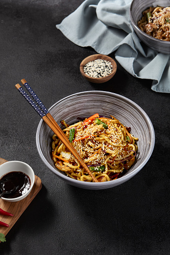 Wheat noodles with a variety of fresh vegetables, presented in a grey bowl, accessorized with chopsticks, on a dark backdrop. Sauce and other ingredients are scattered for additional appeal.