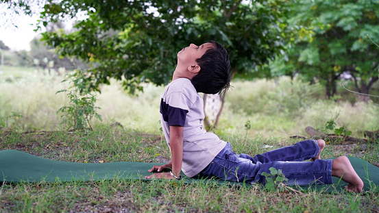 A little boy with long hair does yoga at garden. Teenager sits in lotus position on the outdoor. Happy little boy exercising sport at home