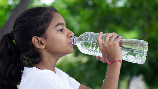 Happy Cute Indian girl with black hair drinking water in the park, holding plastic bottle and showing thumb up. outdoor shot