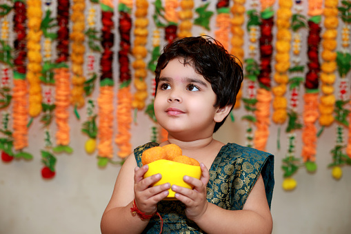 3-4 years baby girl holding sweet in bowl against floral background on Diwali festival.
