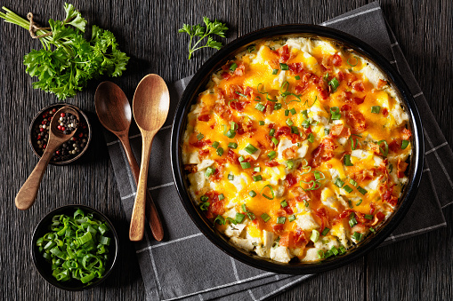 loaded up chicken bake of biscuits layered with a cooked chicken meat in creamy sauce and topped with green onions, cheese, and bacon in baking dish on dark wooden table, flat lay