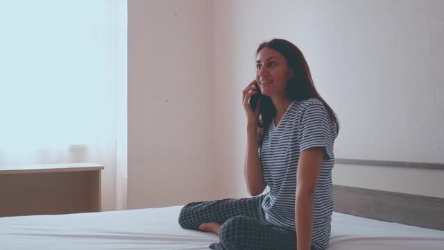 Brunette woman sitting in white bed using smartphone, having video call, smiling young woman relaxing in bedroom, waking up in the morning and sending text messages on one device, technology concept