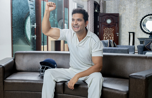 Overjoyed man in cricket dress celebrating win sitting on sofa at living room, Man enjoying watching cricket at home