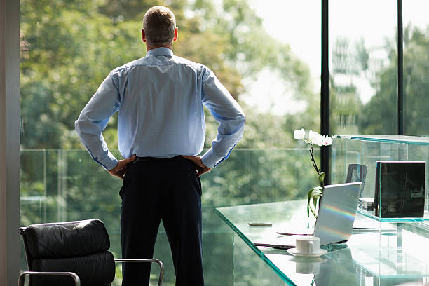 empresario mirando por la ventana de oficina - mano sobre la cadera fotografías e imágenes de stock