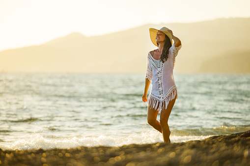 Young happy woman with sun hat taking a walk in summer day on the beach. Copy space.