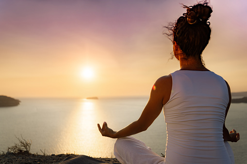 Back view of a relaxed woman doing Yoga meditation exercises in Lotus position on a hill above the sea at sunrise. Copy space.