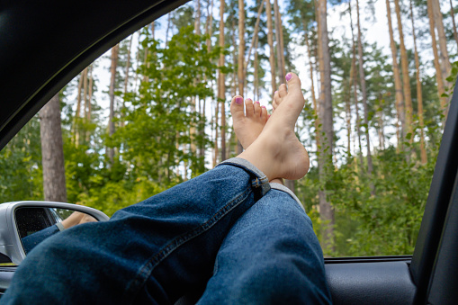Female feet in blue jeans out of car window. Concept of comfortable travel vacation holiday. Getting away to make real candid moments. Reduce carbon footprint. Woman resting in road trip refresh and recharge