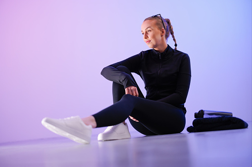 Thoughtful young woman in black sportswear sitting by bottle against purple background