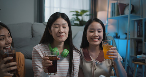 Selective focus Asian young woman and her friend clink a glass of beverage and drinking while party in living room at home, They are happiness during celebration of friendship