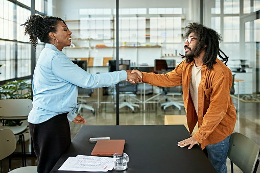 Side view of mature Black woman and multiracial bearded man, eye to eye and shaking hands after their time together in see-through conference room.